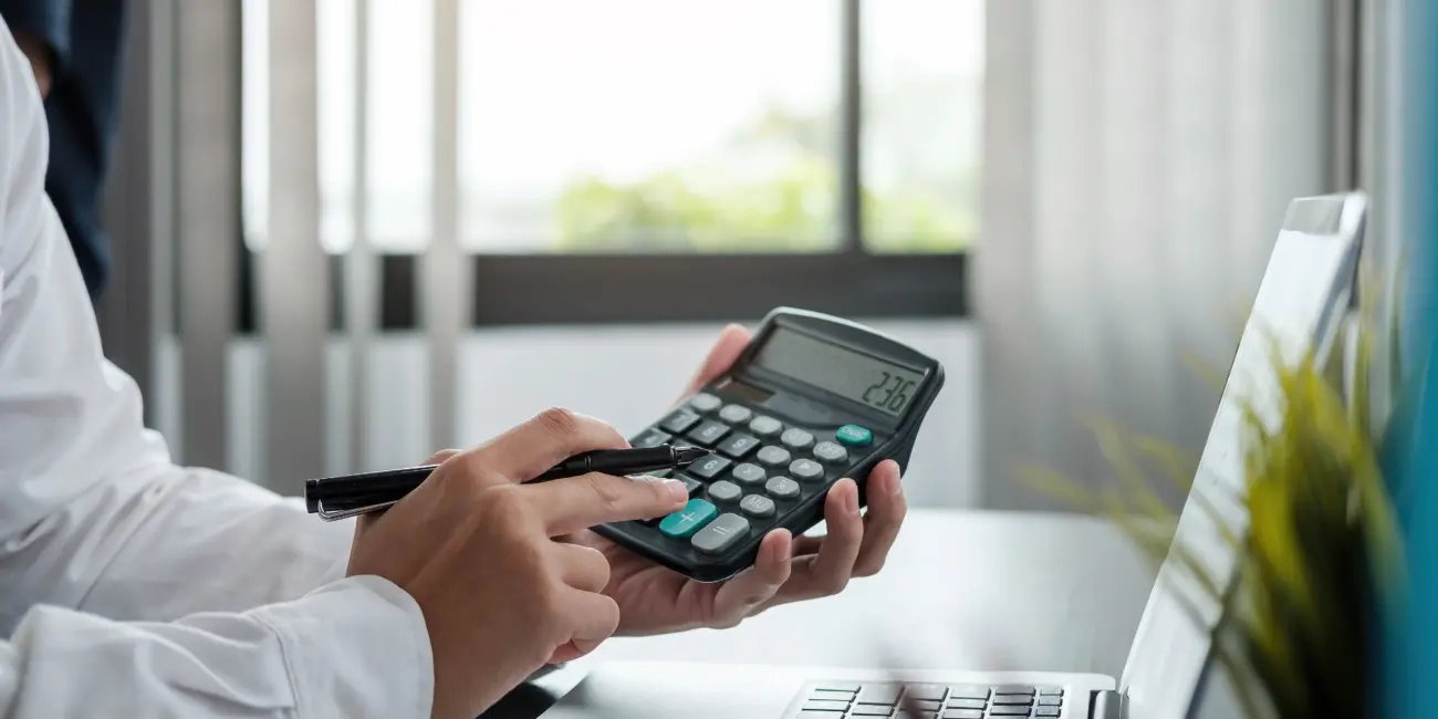 Photo of an accounting sitting in front of a laptop and using a handheld calculator while holding a pen